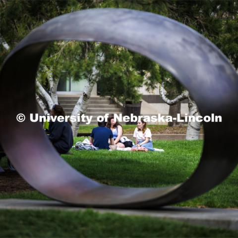 Framed by the sculpture Fragment X-O, Paige Vose, center, Makena Niehaus and Evan Mott (back to camera) enjoy the sunshine and awesome weather at the Sheldon sculpture garden. April 17, 2024. Photo by Craig Chandler / University Communication and Marketing.