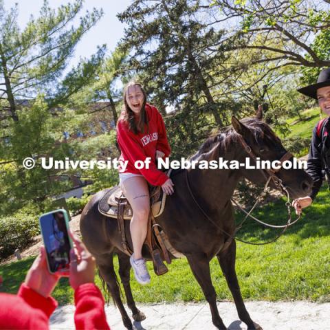 Kyrnlie Simpson laughs as she sits on a horse as her friend, Ophelie Wilson takes a photo of her. Simpson said “this was the best thing ever” for her. The Nebraska Rodeo Team gave Huskers a chance to be up close to their horses and even go for a ride. The team filled the space northwest of the Union to help promote their Nebraska Cornhusker College Rodeo being held Friday and Saturday. April 29, 2024. Photo by Craig Chandler / University Communication and Marketing.