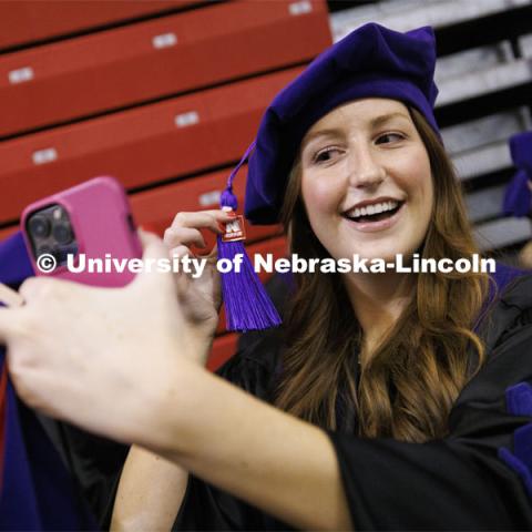 Emilee Smith uses her phone as a mirror to adjust her tassel so the logo shows. College of Law commencement in Devaney on the volleyball court. May 3, 2024. Photo by Craig Chandler / University Communication and Marketing.