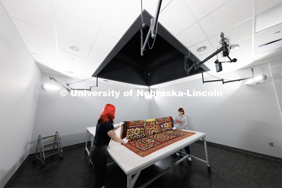 Student workers Olivia Fiore, left, and Inas Hskan, fold a quilt after it was photographed.  The quilt is titled “Soldier’s Quilt” c. 1850-1910 and probably made in India. The area above the quilt leads to a second-floor balcony with a camera suspended above the table for photographs. Quilt Center. February 8, 2023. Photo by Craig Chandler / University Communication.