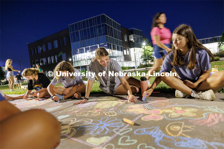 Brooke Poppert, a senior from St. Paul, Nebraska, center, chalks artwork on the sidewalk across from Cather Dining Complex. The Salt Company, a local college ministry, held a gathering Wednesday night outside Cather Dining Complex for fellowship, games and chalk painting on the sidewalk. August 16, 2023. Photo by Craig Chandler/ University Communication.