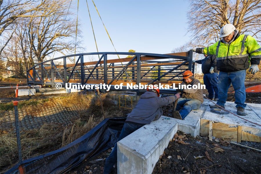 Workers with Judds Brothers Construction adjust a long bolt so it aligns with the bridge as it is lowered onto the bolts on the east side of the creek. A new footbridge over Dead Mans Run on East Campus’ Maxwell Arboretum. December 5, 2023. Photo by Craig Chandler / University Communication and Marketing.