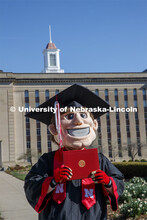 Herbie Husker stands in front of Love Library, proudly holding a diploma and is decked out in gradua