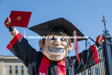 Herbie Husker stands in front of Love Library, proudly holding a diploma and is decked out in gradua