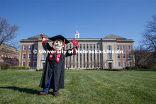 Herbie Husker stands in front of Love Library, proudly holding a diploma and is decked out in gradua