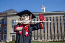 Herbie Husker stands in front of Love Library, proudly holding a diploma and is decked out in gradua
