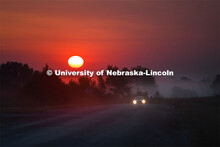 A car drives up out of the fog on a rural Nebraska road as the sun rises in the early morning sky. S